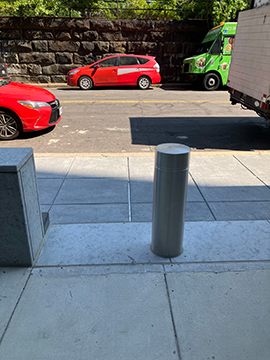 A picture of a pathway to a sidewalk and a street. In the foreground, there is the end of a half wall on the left and a bollard slightly to the right of center of the picture. Beyond that in the background, there are two red taxis in the street as well as two trucks, all of which are parked on the side of the street. There is a sidewalk between the pathway immediately in the foreground and the street in the background.